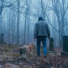 Ich sehe keinen Text. Eine Person mit Kapuze und Hund geht durch einen nebligen Friedhof im Wald.