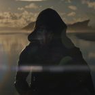 Ein düster gekleideter, maskierter Charakter steht am Strand bei Sonnenuntergang, reflektiert im Wasser.