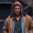 Ein Mann im braunen Mantel und rotem Hoodie steht entschlossen vor einer dunklen Treppe.