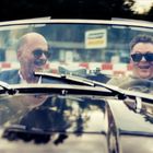 Zwei Männer mit Sonnenbrillen in einem offenen Auto, lachend. Im Hintergrund eine Rennstrecke mit rot-weißen Absperrungen.