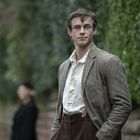 Ein Mann in brauner Jacke und weißem Hemd steht draußen, unscharfe Person im Hintergrund; grüne Hecke und Mauer.