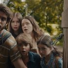 Fünf Personen blicken neugierig und überrascht durch einen Spalt in einer Holztür, umgeben von grüner Vegetation.