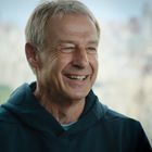 Eine lächelnde Person in einem Hoodie vor einer verschwommenen Stadtlandschaft im Hintergrund.