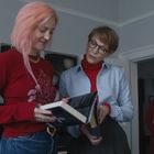Zwei Personen lesen ein Buch, eine hat rosa Haare. Im Hintergrund steht eine ältere Person und ein Klavier ist zu sehen.