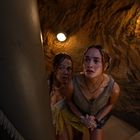 Zwei junge Frauen, nass und ängstlich, stehen auf einer Wendeltreppe in einer dimm beleuchteten Umgebung.