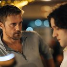 Zwei Männer in einem Gespräch, einer fokussiert, der andere unscharf, mit verschwommenen Lichtern im Hintergrund.