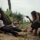 Ein Mann sitzt im Sand, eine Frau hält ihm ein Messer entgegen. Umgeben von tropischer Vegetation und einem provisorischen Unterschlupf.