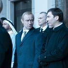 Mehrere Männer stehen formell gekleidet draußen und blicken ernst; einige tragen Anzüge, einer hat traditionelle arabische Kleidung.