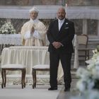 Ein Priester und ein Mann im Anzug stehen in einer geschmückten Kirche, flankiert von Blumenarrangements.