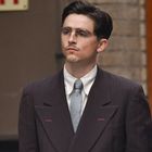 Ein "EXIT"-Schild ist im Hintergrund. Ein Mann im Anzug mit Krawatte, Brille und kleinem Bart schaut ernst.