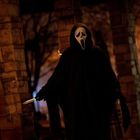 Eine Figur in schwarzem Umhang mit weißer Maske und Messer steht zwischen Ziegel- und Steinbögen in dunkler Umgebung.