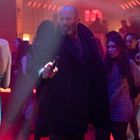 Leuchtreklame „ND“, Mann mit Mantel und Pistole in lebhafter Clubszene, viele tanzende Menschen, bunte Lichter.