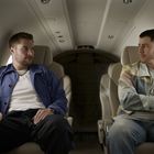 Zwei Männer sitzen in einem Privatjet, sie unterhalten sich. Die Sitze sind beige, und die Beleuchtung ist gedämpft.