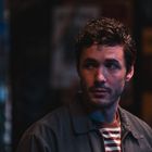 Ich kann den Text nicht erkennen. Ein Mann in einem gestreiften Shirt und Jacke schaut zur Seite in einem dunklen Raum.