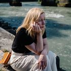 Eine Frau in schwarzem Shirt und heller Hose sitzt nachdenklich an einem Fluss.