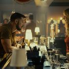 Ein Mann und eine Frau stehen an einer Bar. Er lehnt sich vor, sie hört aufmerksam zu. Die Bar ist voll beleuchtet.