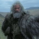 Ein alter Mann mit langem grauem Haar und Bart in einem Fellumhang steht in einer kargen, hügeligen Landschaft.