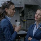 Zwei Frauen in blauen Uniformen plaudern in einer Flugzeugküche. Eine hält einen Pappbecher mit einem grünen Logo.