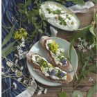 "April" in roter Schrift. Zweimal Baguette mit Frischkäse, Blumen, Wildkräuter und Dip auf einem Holztisch.