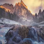 1 • 2027. Ein Berg wird im goldenen Licht der untergehenden Sonne beleuchtet, im Vordergrund ein fließender Bach.