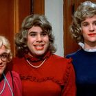 "Keep It Up" steht auf einem kleinen Aufkleber an der Holzwand. Drei Personen tragen Frauenkleidung und Perücken.