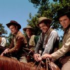 Fünf Cowboys auf Pferden, alle tragen Hüte und Westernkleidung, unter blauem Himmel mit Bäumen im Hintergrund.