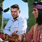 Zwei Männer in traditioneller Kleidung halten einen Mann fest, umgeben von Kakteen in einer Wüstenlandschaft.
