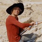 Eine Person mit schwarzem Hut und rotem Pullover hält ein Gewehr in einer sandigen Landschaft.