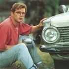 Ein Mann mit Brille repariert ein altes Auto, hält ein Werkzeug. Trägt rotes Shirt und Jeans, sitzt auf dem Boden.