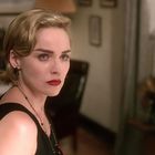 Eine Frau mit kurzen blonden Haaren und rotem Lippenstift blickt ernst. Im Hintergrund ist ein verschwommenes Zimmer zu sehen.