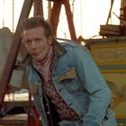 Ein Mann in Jeansjacke und rosa Schal schaut in die Kamera, umgeben von Jahrmarkt-Dekor und einer Leiter im Hintergrund.