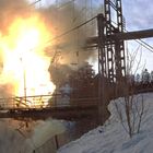 Starke Explosion auf einer Brücke in verschneiter Landschaft; Funken und Trümmer fliegen umher.