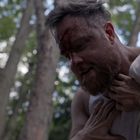 Ein Mann im Unterhemd mit blutverschmiertem Gesicht, umgeben von Wald, wird am Kragen gepackt.