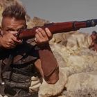 Ein Mann in postapokalyptischer Kleidung zielt mit einem Gewehr in einer felsigen, wüstenartigen Landschaft.