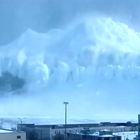 Eine riesige Welle aus Schnee oder Nebel rollt über ein Gebäude und einen Kran. Die Umgebung wirkt winterlich und kalt.
