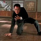 Ein Mann kniet erschöpft auf einem Holzboden in einem Kampfraum. Er trägt Anzug, Blut an der Stirn. Im Hintergrund japanisches Dekor.