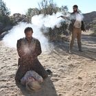 Ein Mann schießt auf einen knienden Mann, der über einer am Boden liegenden Person kauert, in einer wüstenartigen Landschaft.