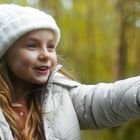 Ein lachendes Mädchen im Wald zeigt mit dem Finger. Sie trägt eine weiße Mütze und eine Jacke.