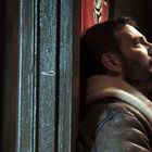 Ein Mann mit Bart lehnt nachdenklich an einer dunklen Holzoberfläche und hält ein Gewehr.