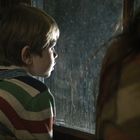 Ein Kind mit einem gestreiften Pullover blickt aus einem Fenster. Es sieht nachdenklich aus.