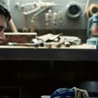 Ein Mann sitzt in einer Werkstatt und blickt auf ein blaues, fremdartiges Wesen. Im Hintergrund sind Werkzeuge zu sehen.