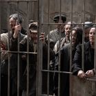 Mehrere Menschen hinter rostigen Metallgittern, teils mit besorgten Gesichtern, tragen abgetragene Kleidung.