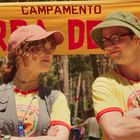 Text: "CAMPAMENTO GARDA DE OSO". Zwei Personen in gelben T-Shirts und Hüten lächeln sich an. Hintergrund aus Bäumen.