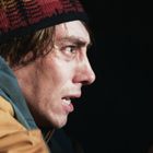 Ein Mann mit entschlossenem Ausdruck trägt eine bunte Mütze und eine orange Jacke in dunkler Umgebung.