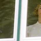 Eine Frau blickt nachdenklich durch ein Fenster. Sie trägt eine weiße Bluse. Draußen sind grüne Bäume.