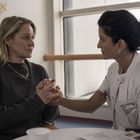 Zwei Frauen halten sich an den Händen. Eine trägt weiße Kleidung, die andere einen grauen Pullover. Im Hintergrund sind bunte Vögel.