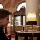 Zwei Personen sitzen in einem eleganten Raum mit Säulen. Eine Tischlampe beleuchtet den Tisch zwischen ihnen.