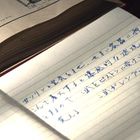 Handschrift auf Papier, geschrieben mit einem Füllfederhalter, daneben technische Zeichnungen in einem Buch.