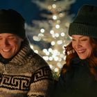 Ein lächelndes Paar in Winterkleidung vor einem unscharfen, leuchtenden Weihnachtsbaum im Hintergrund.