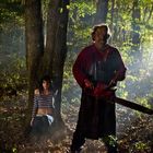 Eine Frau sitzt verängstigt an einen Baum gelehnt, während eine Gestalt mit Kettensäge im Wald steht.